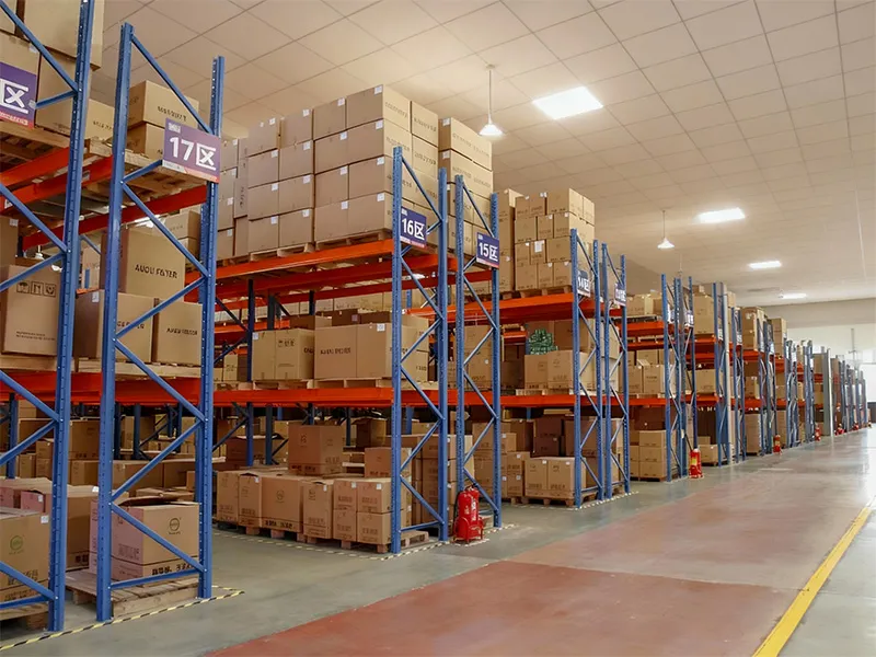 A large-scale industrial warehouse featuring organized high-bay racking systems with labeled sections, containing palletized boxes of finished products ready for global shipment.