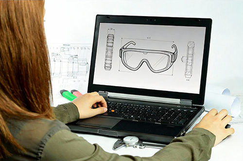 An engineer reviewing technical 2D and 3D drawings of protective eyewear on a laptop, with precision measurement tools and blueprints on the desk, illustrating the product development phase.