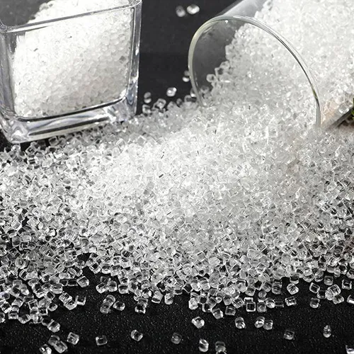 A heap of transparent PMMA (acrylic) plastic granules scattered on a dark background. A filled square glass container stands in the top left, while an overturned cylindrical glass cup on the upper right allows the clear, crystalline resin pellets to pour out and gather in the center, highlighting the high transparency and premium quality of the raw material.