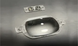 Two high-transparency plastic street light optical lenses displayed on a gray surface. The foreground shows a large rectangular lens featuring an asymmetric curved profiling and flat mounting ears with screw holes on both sides, with a smaller dual-lens module placed in the background.