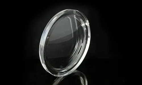 A high-transparency circular plano-convex plastic optical lens, tilted on a reflective black surface and background, showing a thick round outer rim with tiny alignment marks and a smooth convex center surface.