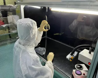 A technician in a full white anti-static cleanroom suit and yellow nitrile gloves performing a visual quality inspection of a transparent circular optical lens under the light inside a laminar flow clean bench in a dust-free workshop.