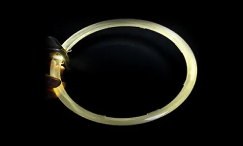 A circular transparent plastic light guide ring undergoing a testing process on a black background, with a hand-held mini LED light source illuminating one end, showing the white light propagating uniformly along the annular rod.