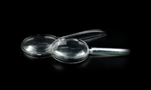 Two handheld magnifying glasses placed overlapping each other on a black background. The front magnifier features a metallic silver handle and a thick biconvex circular lens, while the rear one has an integrated clear plastic handle.