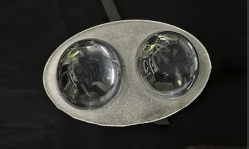 An 8-cavity transparent plastic injection-molded conical optical lens component layout on a black brushed surface, showing a central horizontal runner system that connects eight individual funnel-shaped reflector lens units.