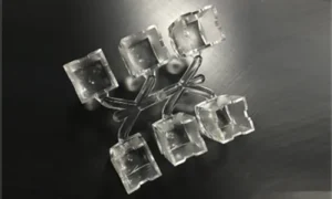 A 6-cavity transparent plastic injection-molded square optical lens component layout on a black brushed surface, showing a central branched runner system that connects six individual hollow square-shaped lens units.