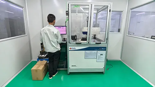 A technician in a cleanroom workshop operating a precision optical measurement instrument with a glass enclosure, monitoring testing data on a computer screen.