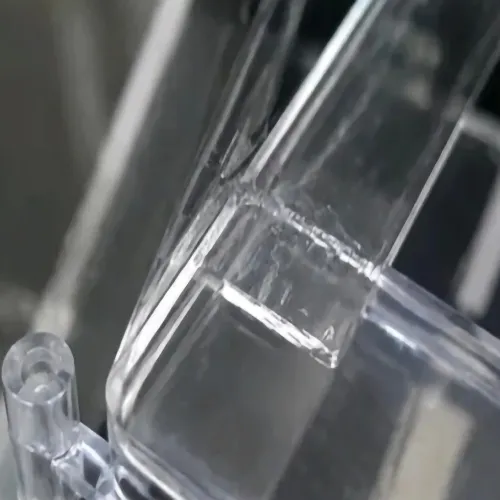 A close-up view of a transparent injection-molded plastic part showing a distinct weld line (knit line) defect. The visible seam formed where two polymer melt fronts met during the filling stage near a structural transition, creating an optical and structural boundary inside the clear PC or PMMA component.