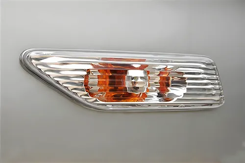 A close-up view of a side indicator repeater lamp assembly integrated into a light gray automotive fender panel. The tricolor component features a clear patterned outer lens with optical fluting and a distinct central amber section with a reflective bezel. The entire unit is framed by a precision-machined chrome rim, highlighting high-quality automotive optics manufacturing via multi-shot injection molding and plating technology.